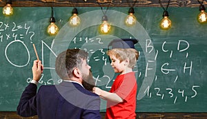 Teacher with beard, father teaches little son in classroom, chalkboard on background. Child in graduate cap listening