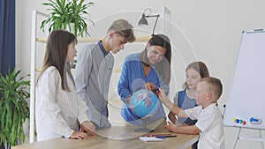 Teacher and children in class are looking at globe, teacher helps explain the lesson to the children in the class at a desk.