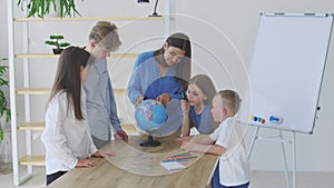 Teacher and children in class are looking at globe, teacher helps explain the lesson to the children in the class at a desk.