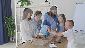 Teacher and children in class are looking at globe, teacher helps explain the lesson to the children in the class at a desk.