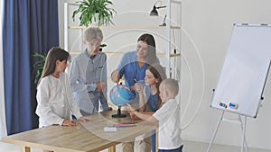 Teacher and children in class are looking at globe, teacher helps explain the lesson to the children in the class at a desk.
