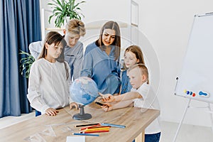 Teacher and children in class are looking at globe, teacher helps explain the lesson to the children in the class at a desk