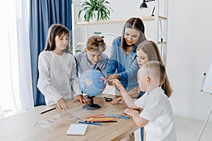 Teacher and children in class are looking at globe, teacher helps explain the lesson to the children in the class at a desk