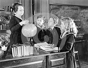 Teacher in a classroom with three pupils pointing to a globe