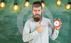 Teacher in eyeglasses holds alarm clock. Lateness concept. Man with beard and mustache on strict face pointing at clock