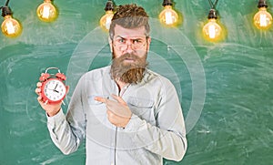 Teacher in eyeglasses holds alarm clock. Lateness concept. Man with beard and mustache on strict face pointing at clock