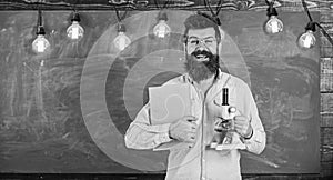 Teacher in eyeglasses holds book and microscope. Science concept. Scientist holds book and microscope, chalkboard on