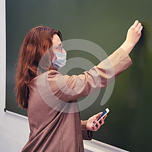 A teacher in a face mask writes a text in chalk on a school blackboard. Concept of problems online school during the coronavirus