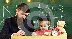 Teacher in formal wear and pupil in mortarboard in classroom, chalkboard on background. Father checking homework, helps