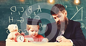 Teacher in formal wear and pupil in mortarboard in classroom, chalkboard on background. Father checking homework, helps