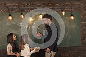 Teacher and girls pupils in classroom near chalkboard. Man with beard in formal suit teaches schoolgirls physics