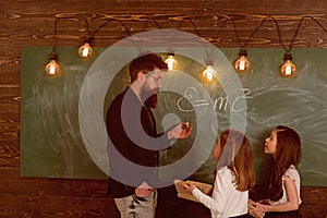 Teacher and girls pupils in classroom near chalkboard. Man with beard in formal suit teaches schoolgirls physics