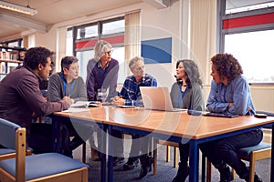 Teacher With Group Of Mature Adult Students In Class Sit Around Table And Work In College Library