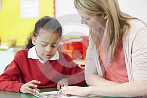 Teacher Helping Female Pupil With Practising Reading At Desk