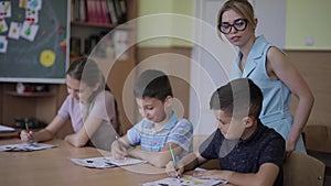 Teacher helping school kids writing test in classroom. education, elementary school, learning and people concept