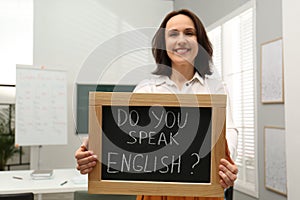 Teacher holding small chalkboard with inscription Do You Speak English? in classroom