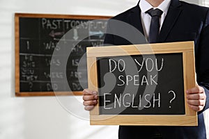 Teacher holding small chalkboard with inscription Do You Speak English? in classroom, closeup