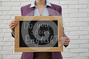 Teacher holding small chalkboard with inscription Do You Speak English? near white brick wall, closeup