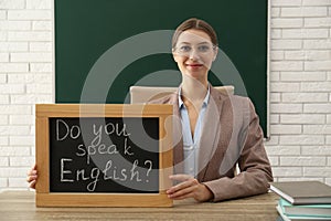 Teacher holding small chalkboard with inscription Do You Speak English? at table in classroom