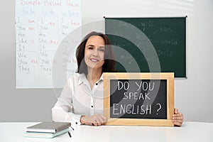 Teacher holding small chalkboard with inscription Do You Speak English? at table in classroom