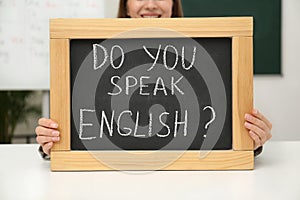 Teacher holding small chalkboard with inscription Do You Speak English? at table in classroom, closeup