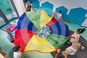 a teacher and kids standing on a circle and holding a multi-colored fabric with many balls on it, nursery creative game