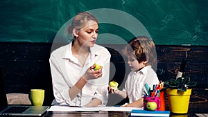 Teacher with a little boy eating apple at a pupil table and counting against a background of a green school board