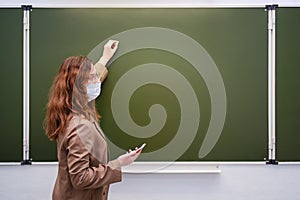 A teacher in a medical mask with a phone in hand writes a text on a school blackboard, copy space. School quarantine education