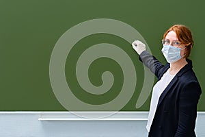A teacher in a medical mask writes with chalk on an empty blackboard, copy space. A woman in a suit stands in a school classroom,