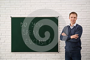 Teacher near green chalkboard with inscription Do You Speak English? in classroom