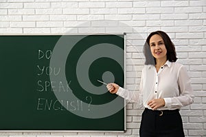 Teacher near green chalkboard with inscription Do You Speak English? in classroom