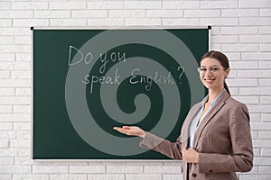 Teacher near green chalkboard with inscription Do You Speak English? in classroom