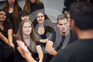 Teacher At Performing Arts School Talking To Students Sitting On Floor In Rehearsal Studio