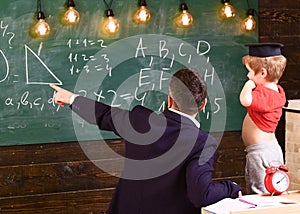 Teacher pointing at the green board while explaining shapes to child. Funny kid in graduation cap pulling up red T-shirt