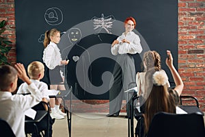 Teacher of primary school teaching to kids anatomy in classroom. Drawing human shape on blackboard. Kids raising hands