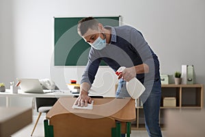 Teacher with protective mask disinfecting desk in classroom. Reopening after Covid-19 quarantine