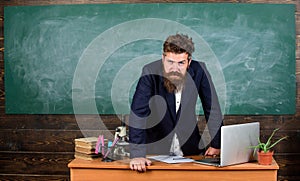 Teacher strict serious bearded man lean on table chalkboard background. Teacher looks threatening. Rules of school