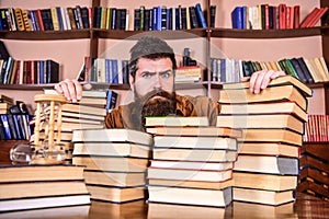 Teacher or student with beard sits at table with books, defocused. Man on serious face between piles of books, while