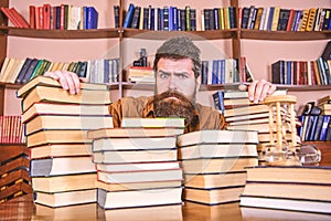 Teacher or student with beard sits at table with books, defocused. Man on serious face between piles of books, while