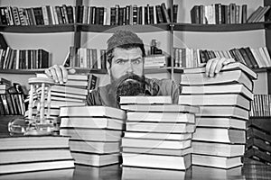 Teacher or student with beard sits at table with books, defocused. Man on serious face between piles of books, while