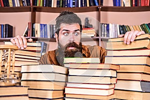 Teacher or student with beard sits at table with books, defocused. Man on shocked face between piles of books, while