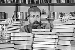 Teacher or student with beard sits at table with books, defocused. Man on shocked face between piles of books, while