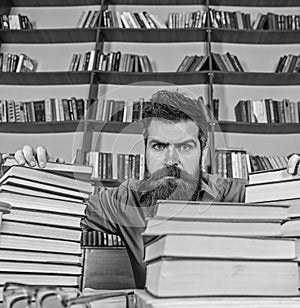 Teacher or student with beard sits at table with glasses, defocused. Man on strict face between piles of books, while