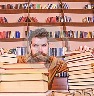 Teacher or student with beard sits at table with glasses, defocused. Man on strict face between piles of books, while