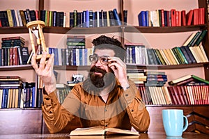 Teacher or student with beard studying in library. Scientist with eyeglasses sits at table and looks at hourglass. Man