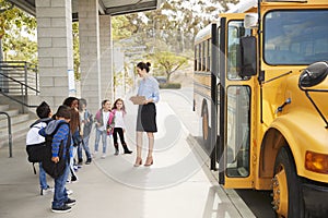 Teacher talks to young school kids by school bus, side view