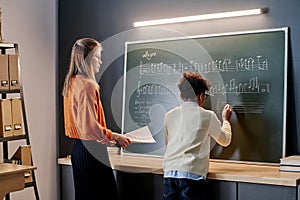 Teacher Watching Her Student Writing Notes On Blackboard