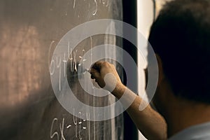 Teacher wearing mask writing equations on a blackboard. Covid situation, pandemic, new normal. Close-up, perspective from the side