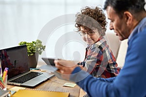 Teaching kid. Portrait of cute little hispanic school boy doing homework together with his father, using technology