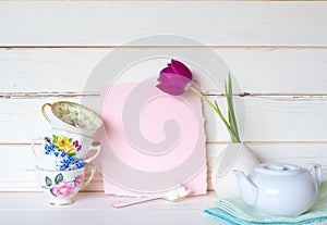 Teacups Stacked with a White Teapot, a Purple Tulip, and Light Pink Invite Card on Table and Against White Board Background with r
